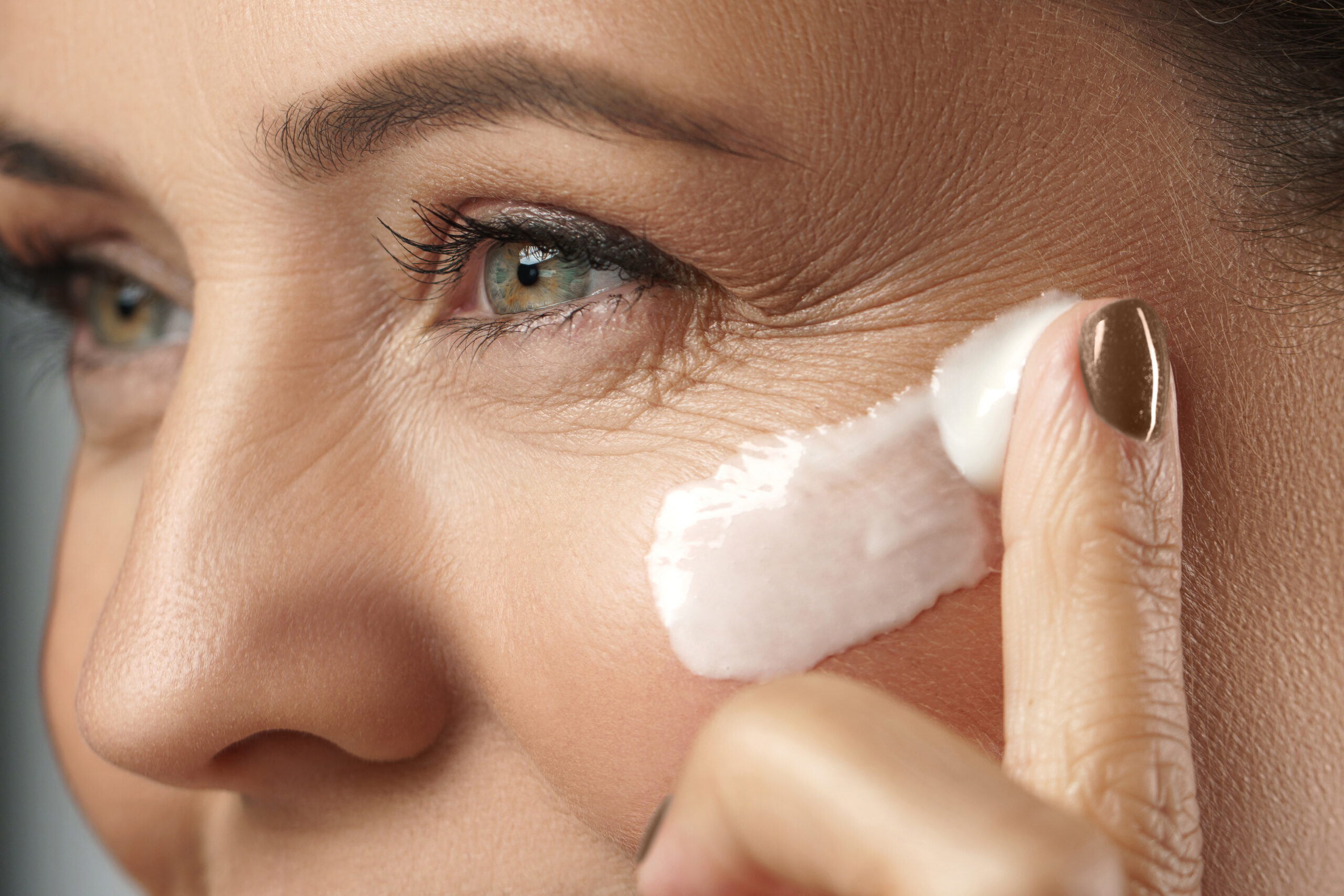 Beautiful middle aged woman applying anti-aging cream on her face on gray