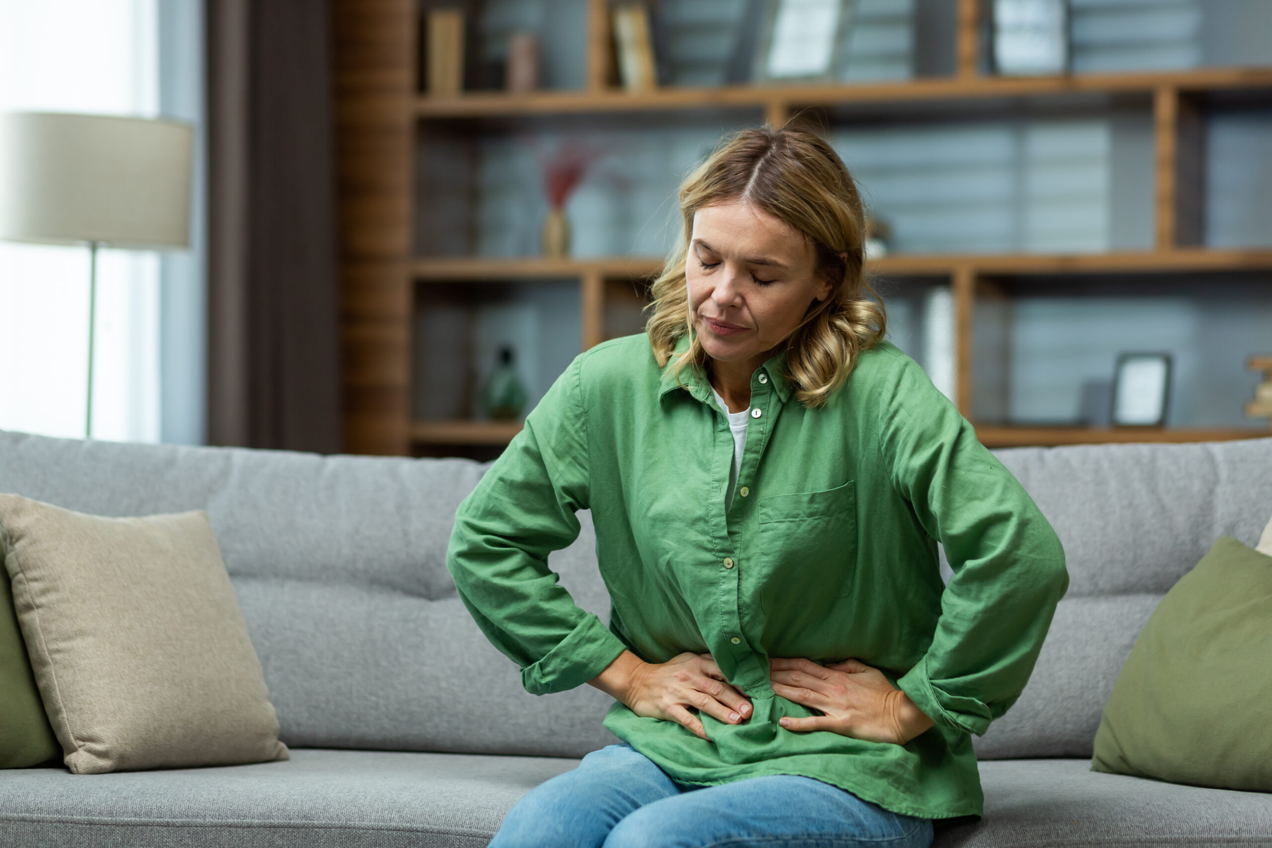 An elderly woman is sitting on the sofa at home, holding her stomach with her hands. Feels the pain of internal organs, indigestion, poisoning, menstrual pain.