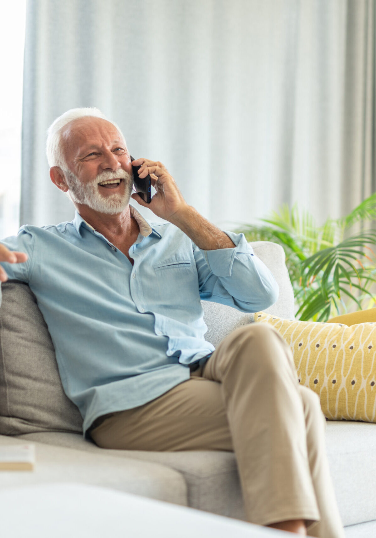 Joyful senior man relaxing on sofa at home while talking on phone and laughing.